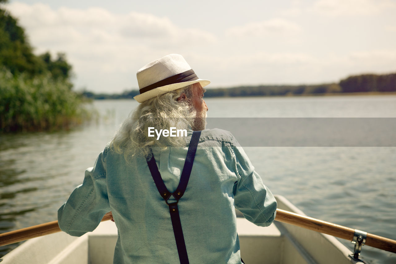 Back view of senior man sitting in rowing boat on a lake wearing suspenders and summer hat