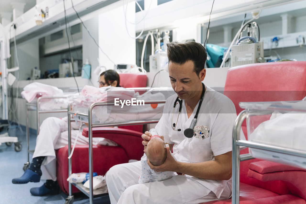 Doctors sitting on armchairs and feeding newborns from bottles holding heads in room with equipment in hospital