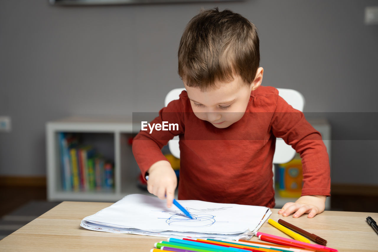 A cute little toddler boy of two years old draws with markers in the album in the children's room