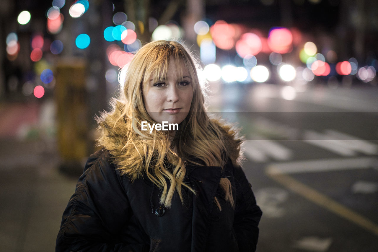 Portrait of young woman standing on sidewalk in illuminated city