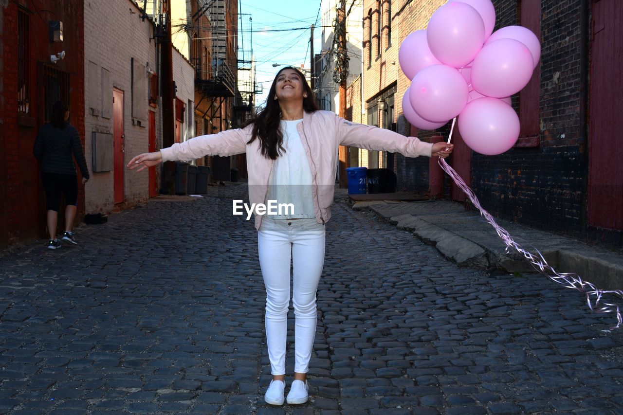 Happy woman holding pink helium balloons while standing on cobbled alley