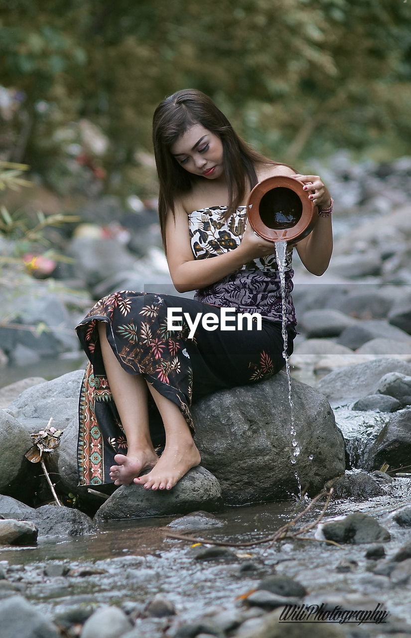 Woman pouring water while sitting on rock in river