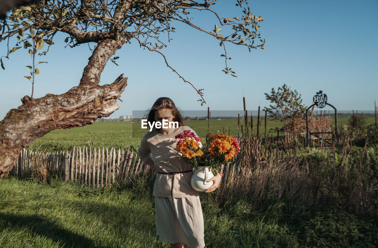 Full length of woman standing on field against sky holding orange flowers