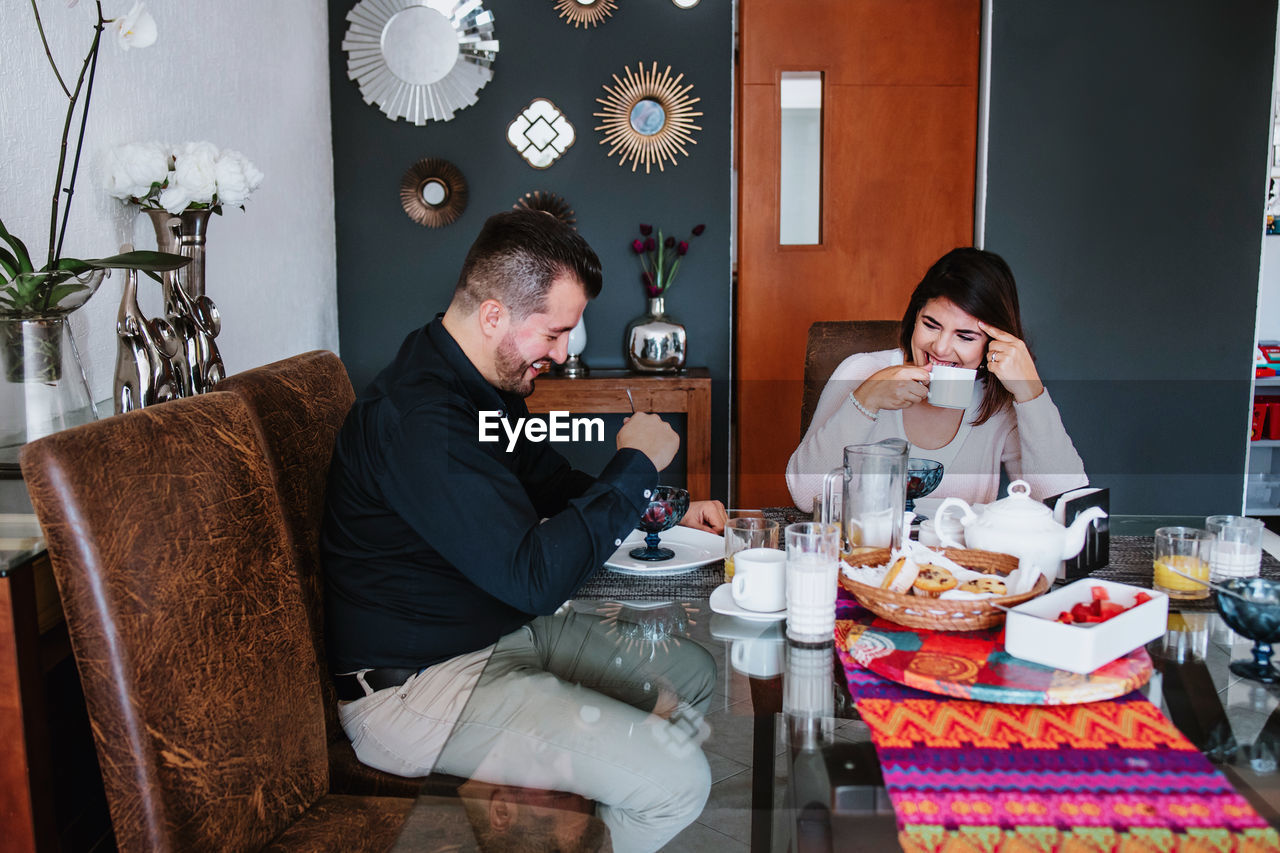 Cheerful young latin american married couple in casual clothes smiling while having breakfast together at home