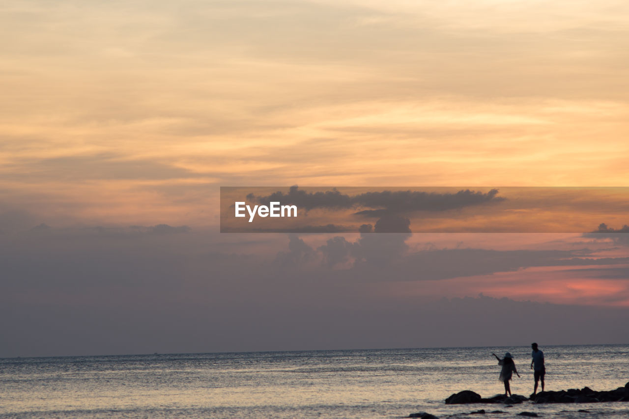 Man and woman on rock by sea against cloudy sky during sunset