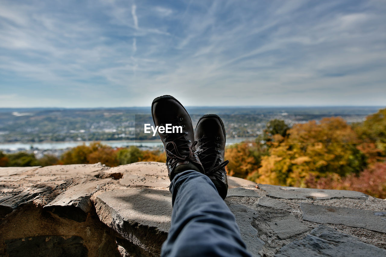 Low section of man relaxing on wall against cloudy sky