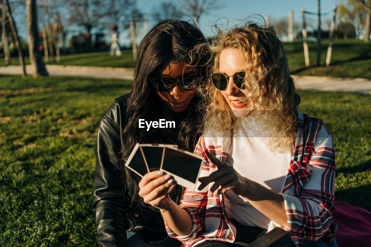 Diverse female best friends relaxing in park and looking through instant photo cards