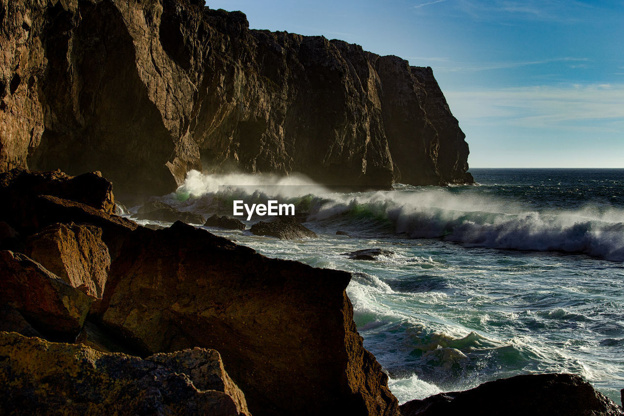 Waves splashing on rocks by sea against sky