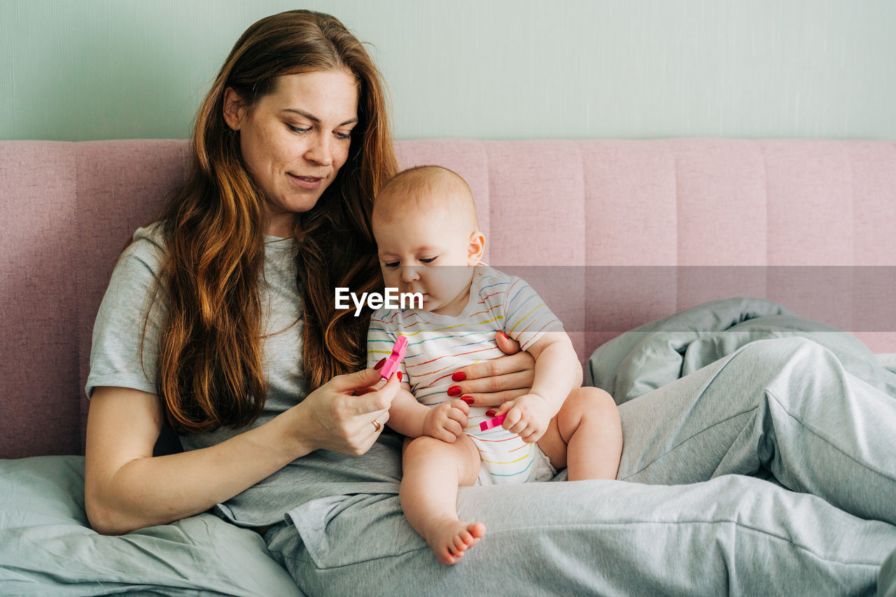 An attractive red-haired mother is sitting in bed, holding the baby in her arms and playing.
