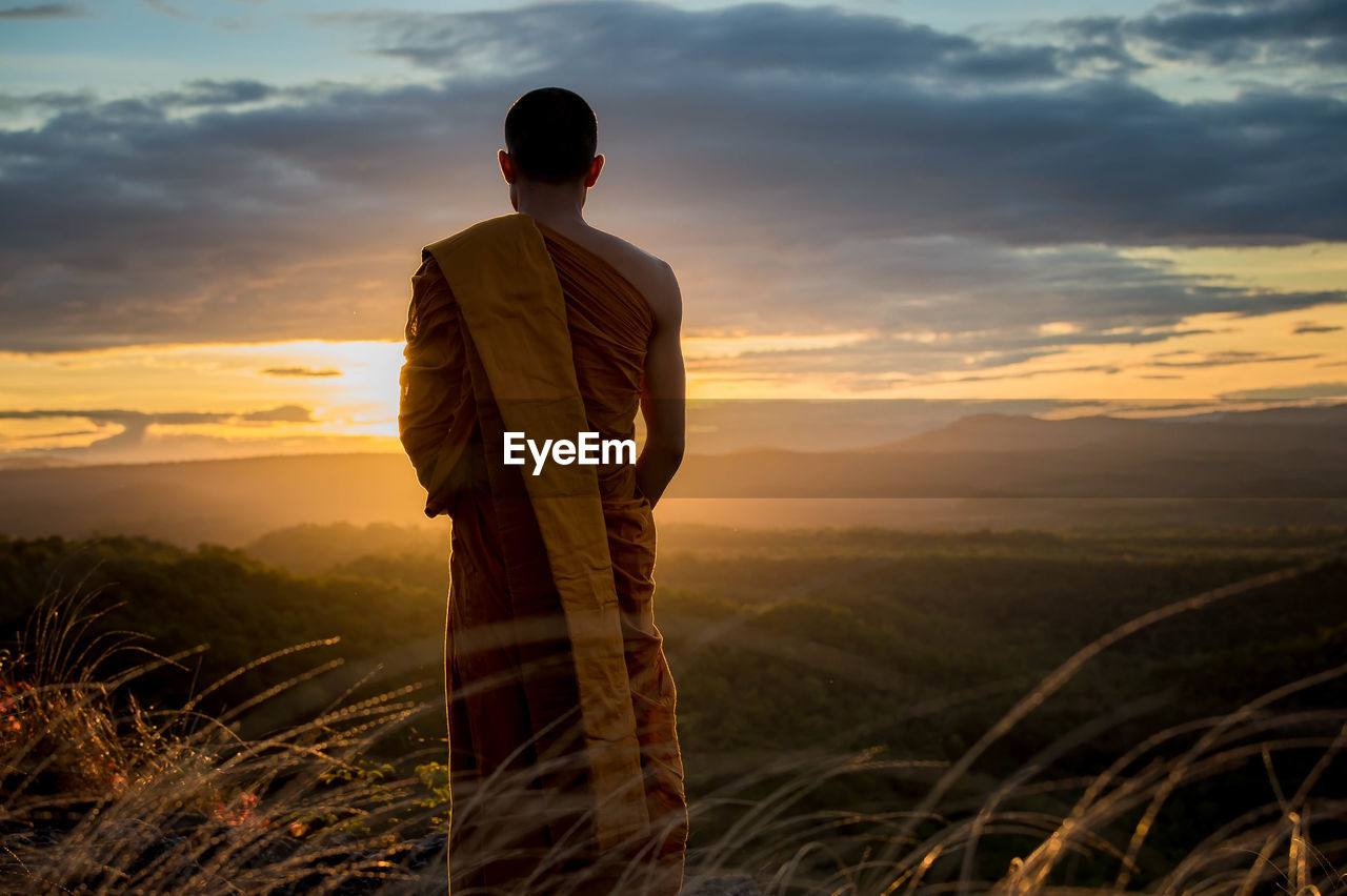 Rear view of monk standing on field against sky at sunset