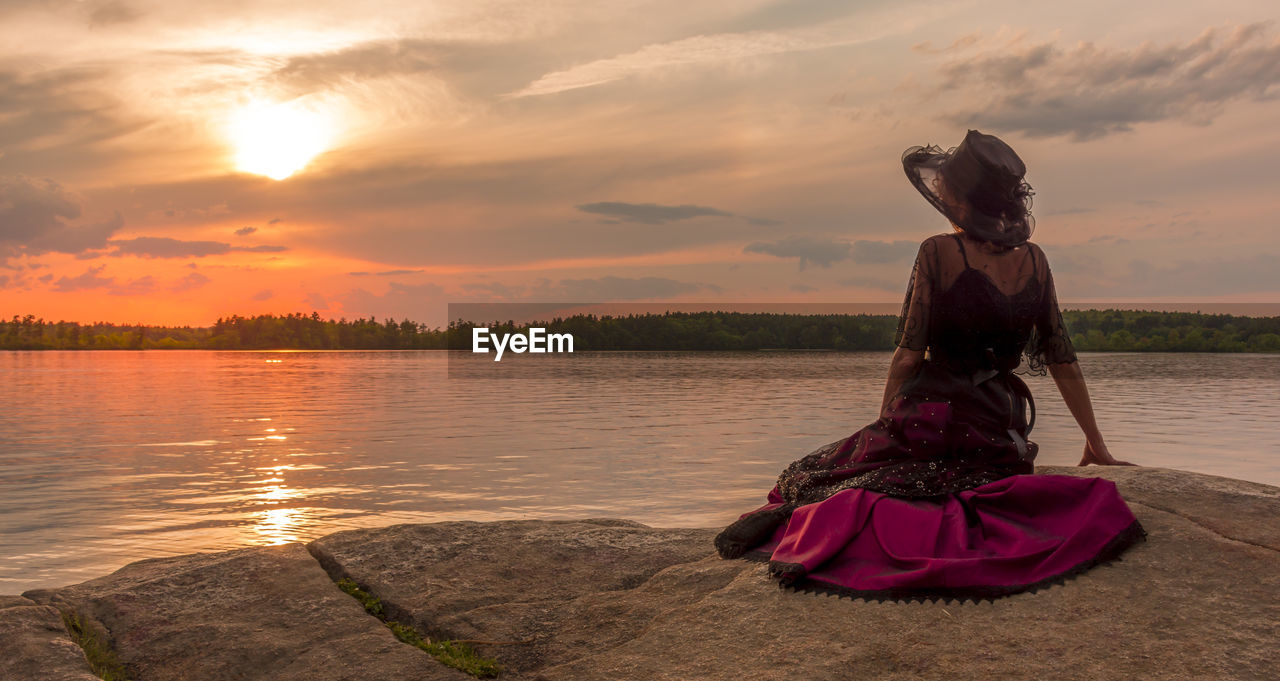 WOMAN SITTING AT LAKE AGAINST SKY DURING SUNSET