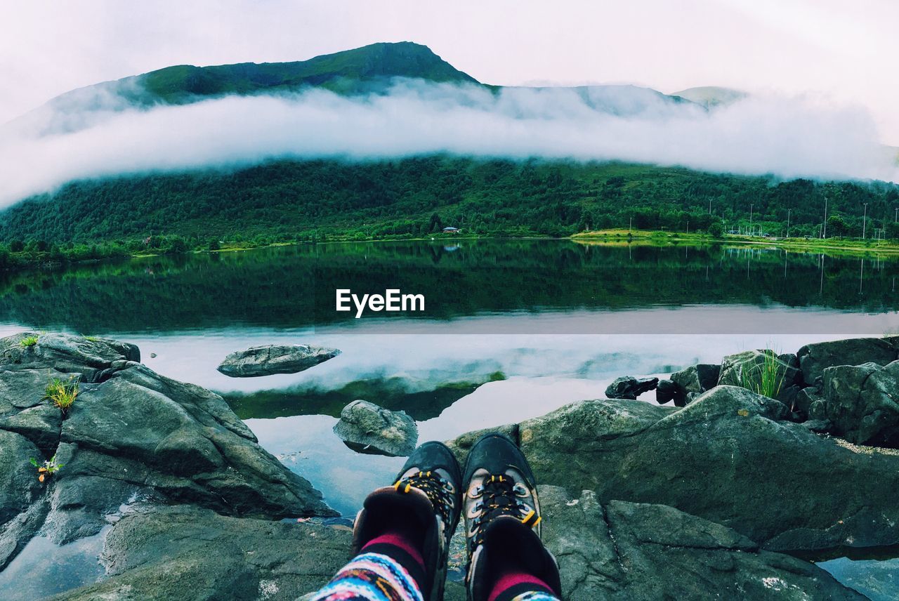 LOW SECTION OF MAN STANDING BY LAKE AGAINST MOUNTAINS