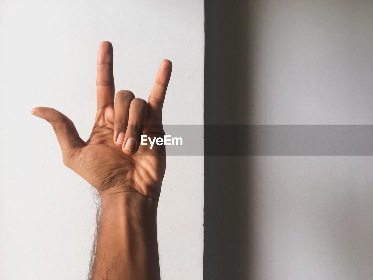 Cropped hand of man gesturing horn sign against wall