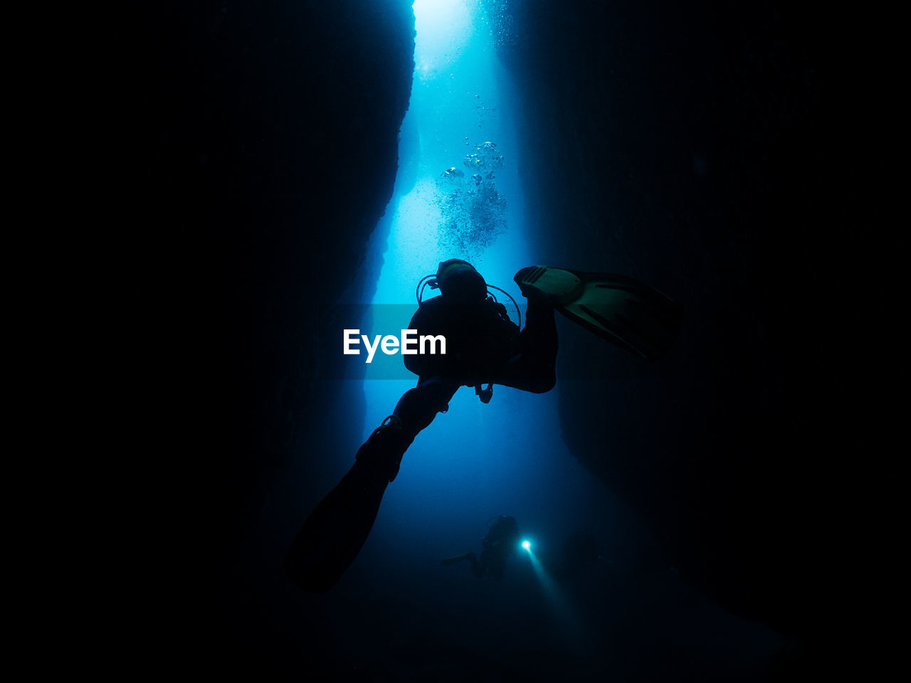Unrecognizable professional divers with scuba and flippers swimming in narrow dark cave with rough stony formations undersea in deep clear water