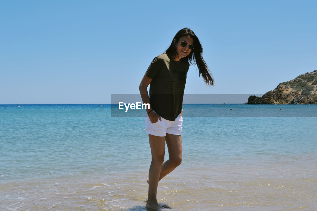 PORTRAIT OF YOUNG WOMAN STANDING ON BEACH AGAINST SKY