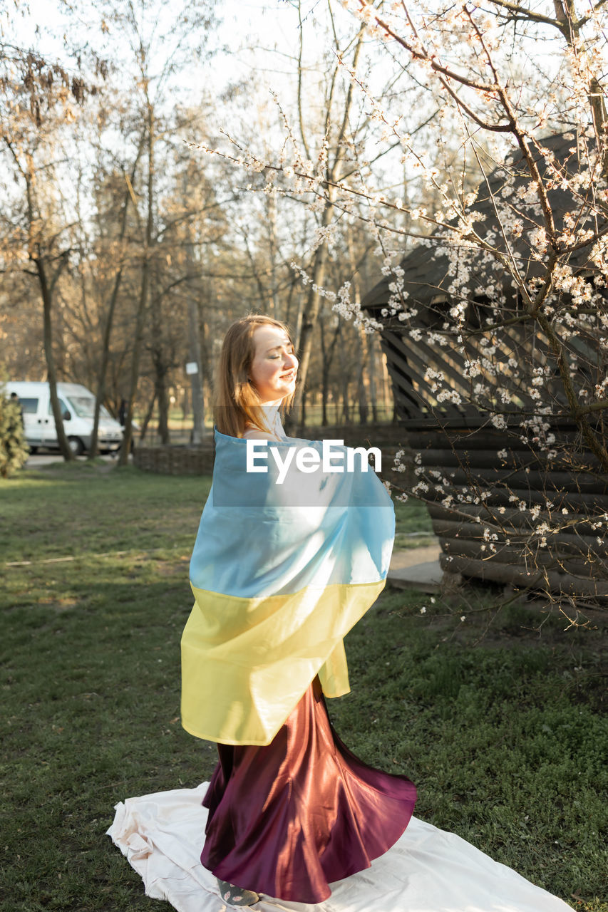 Woman in dress with ukranian flag near trees in spring park