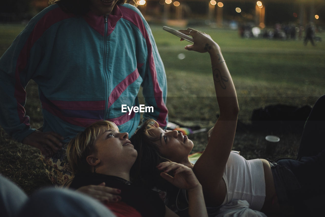 Young woman taking selfie with non-binary person while lying on friend's lap during sunset