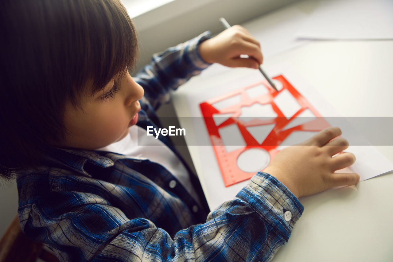 Boy child draws on paper with a ruler on a table sitting by the window