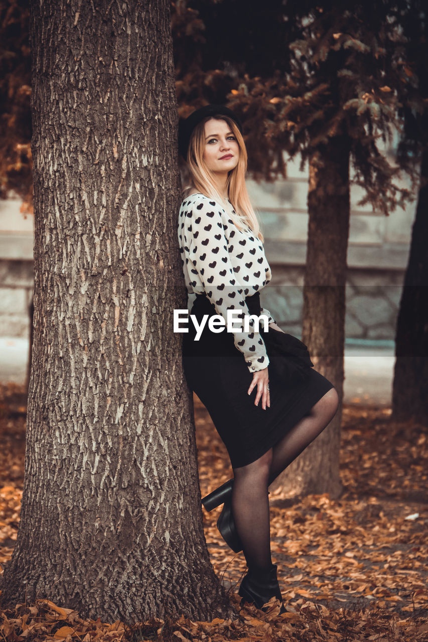 Portrait of smiling young woman standing by tree at public park during autumn