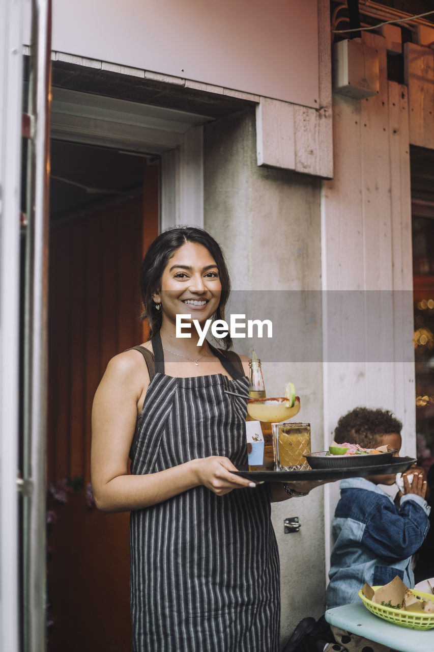 Portrait of smiling waitress wearing apron holding tray of food and drinks at bar