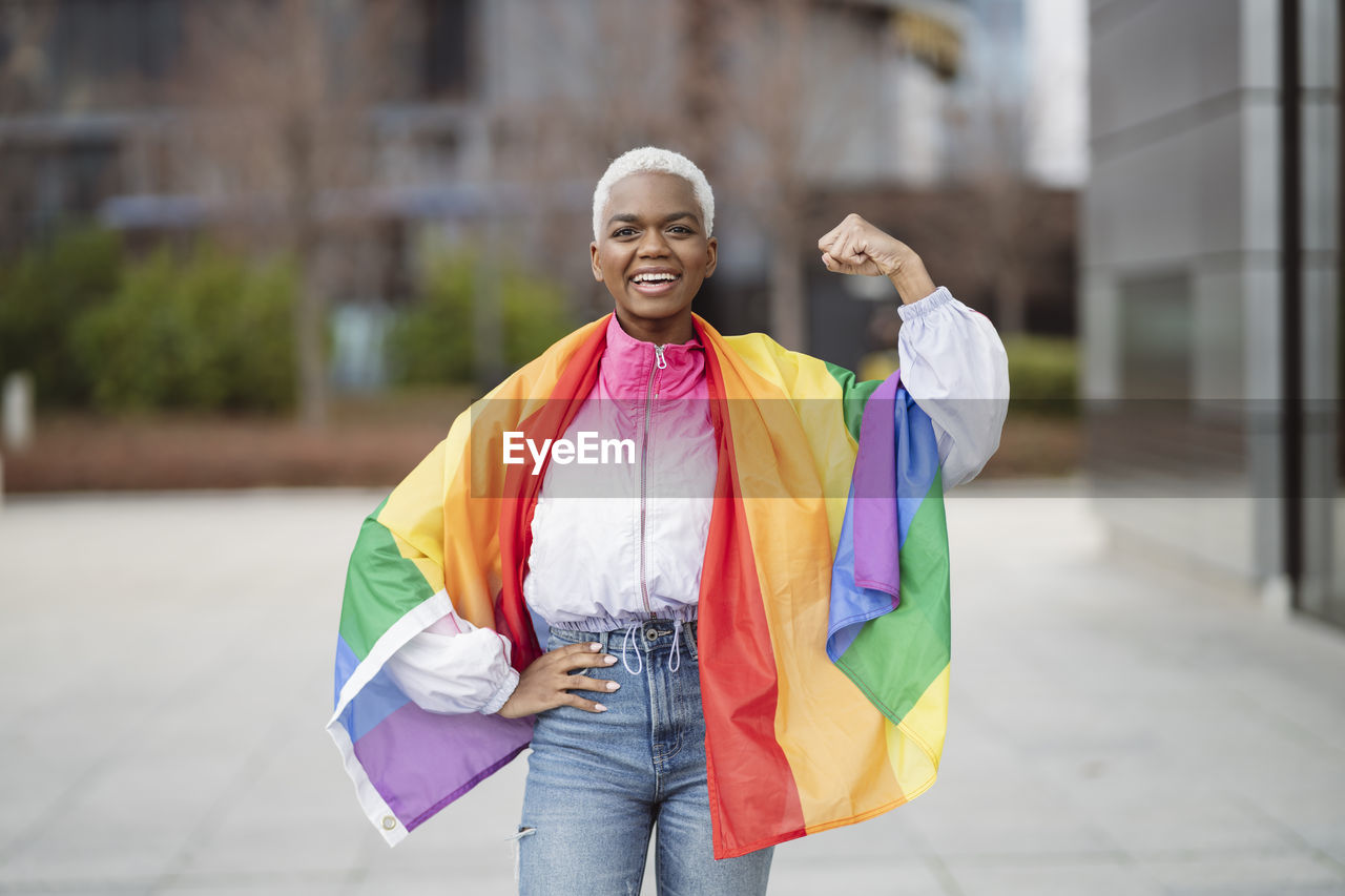 Young woman wrapped in rainbow flag flexing muscle with hand on hip