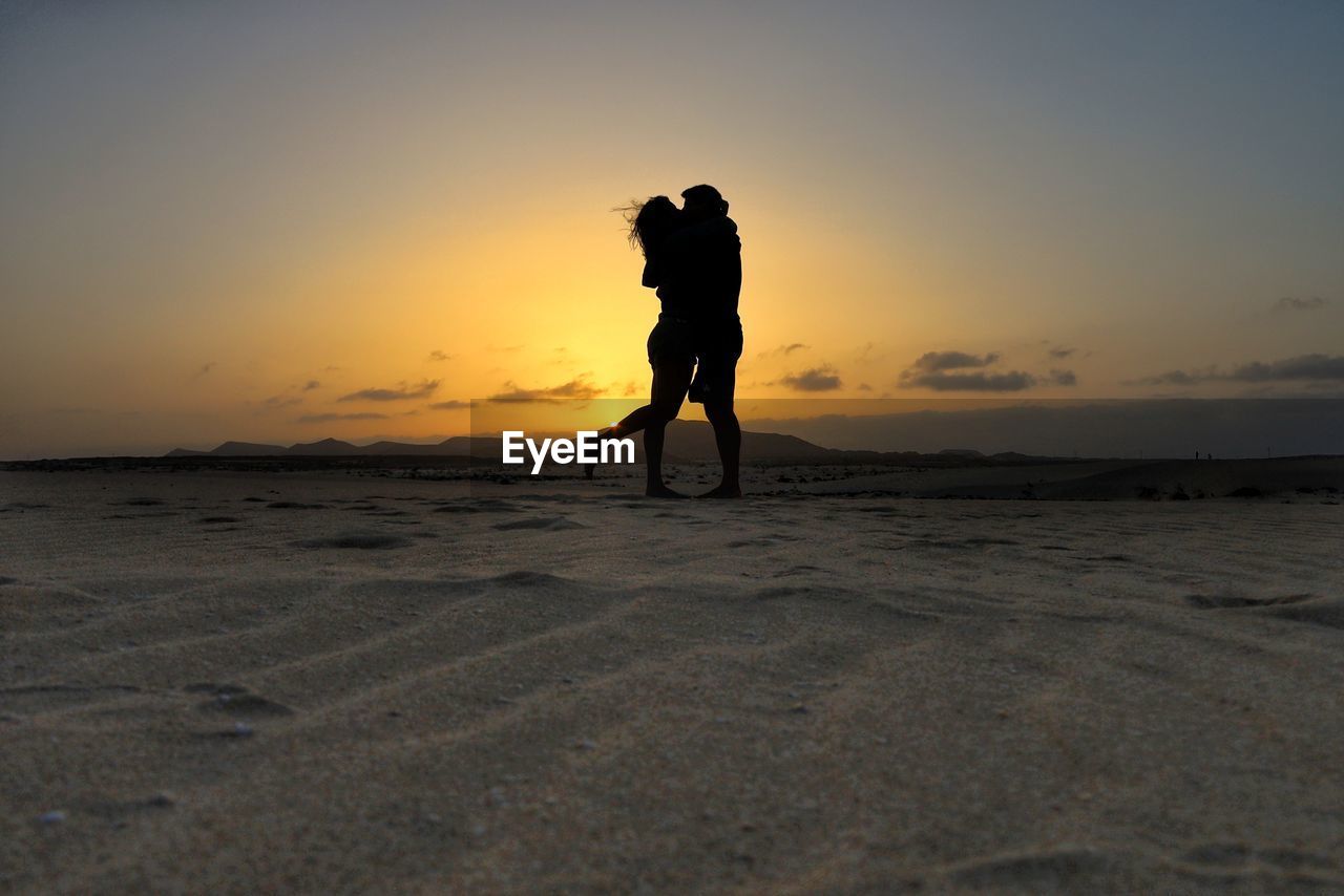 Silhouette couple standing on sand dune against sky during sunset