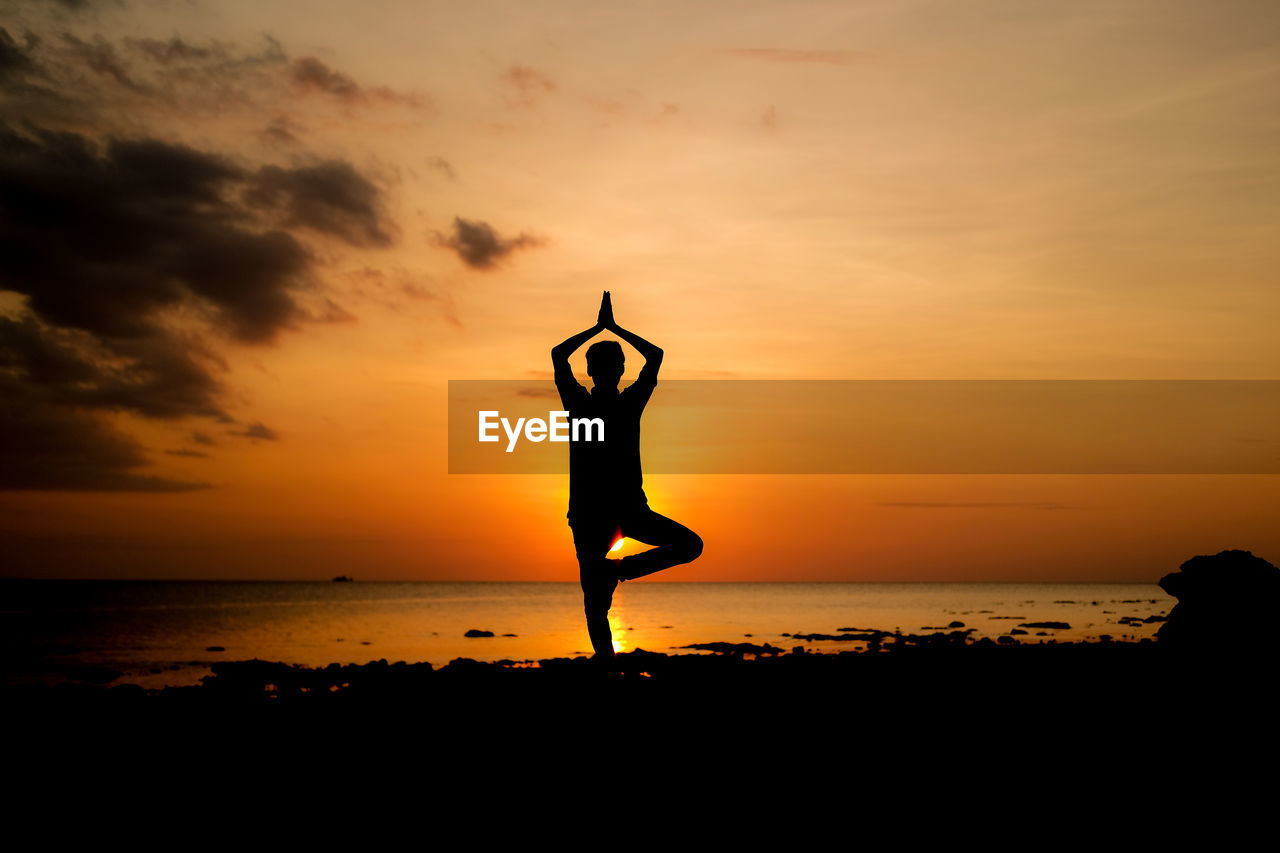 Silhouette person exercising yoga at beach against sky during sunset