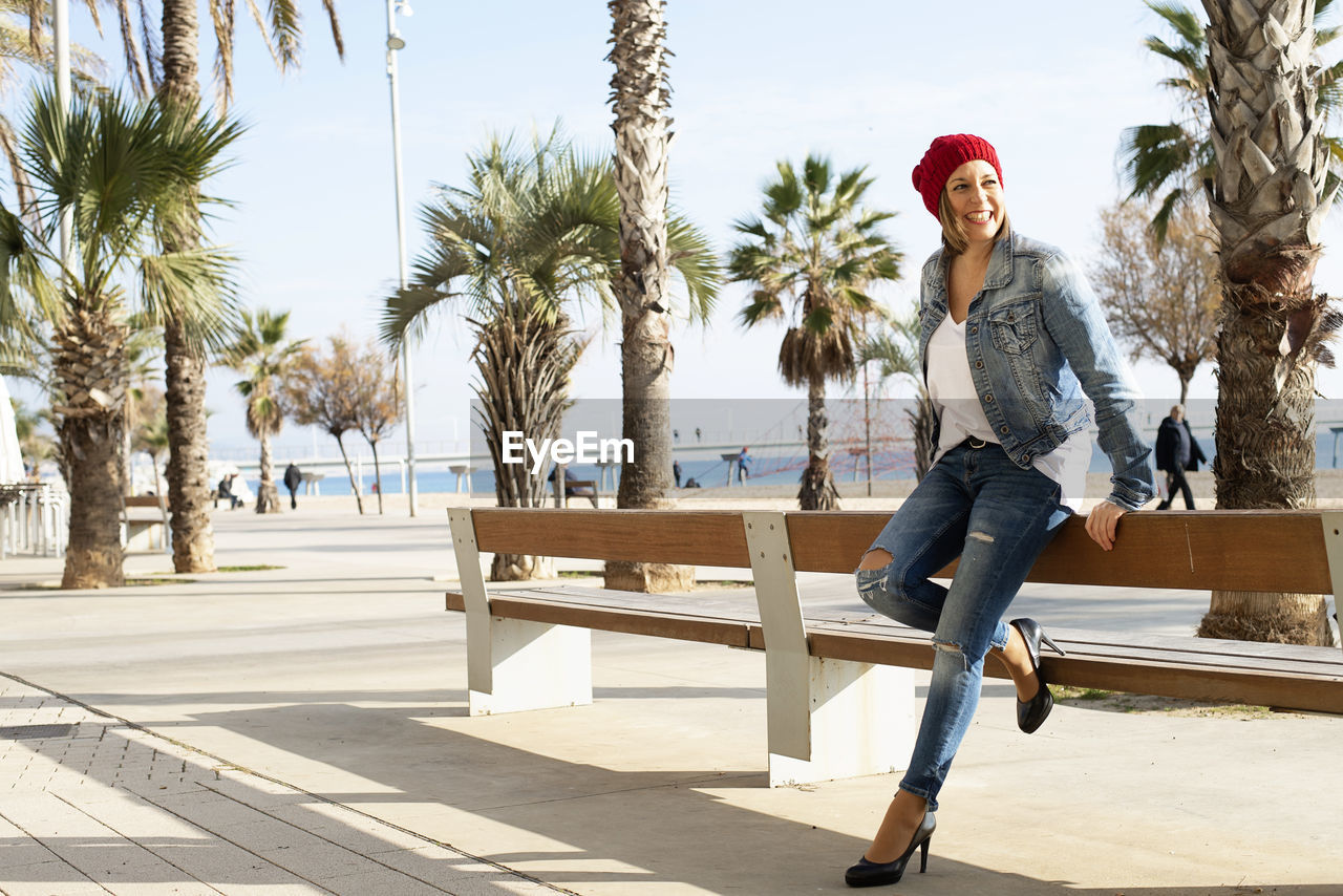 Smiling woman with wool hat leaning on bench in promenade,looking away