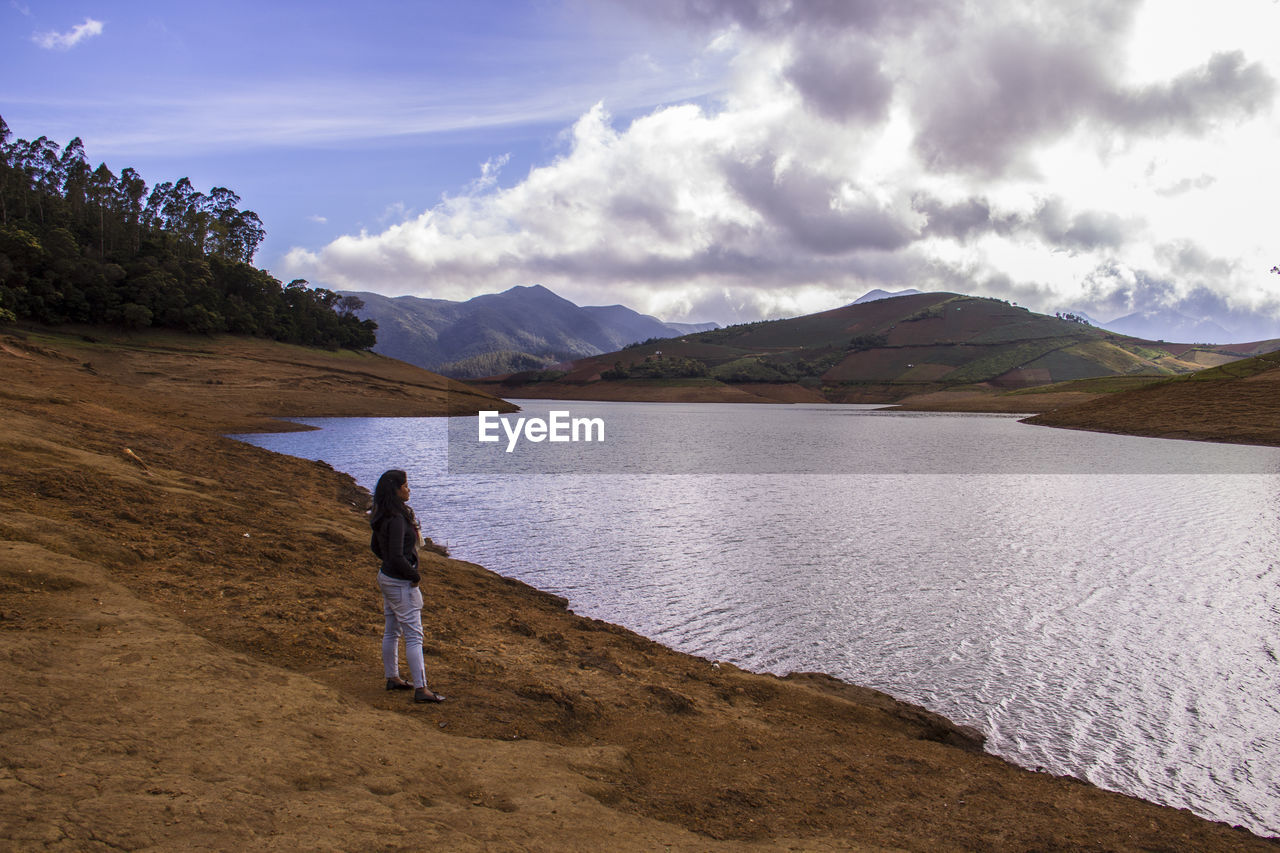 Side view of woman standing at lakeshore against mountains