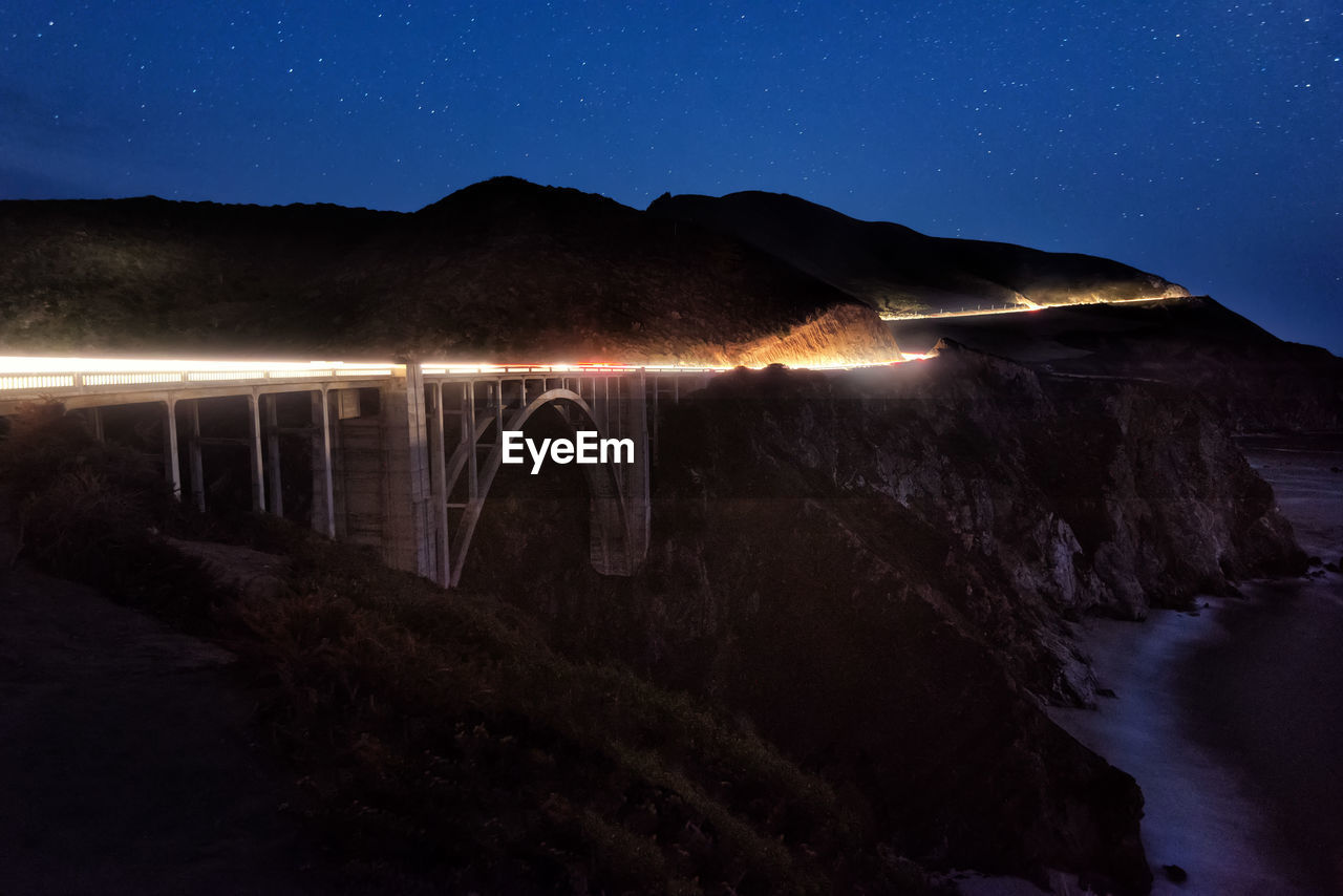 Illuminated bridge over mountains against sky at night