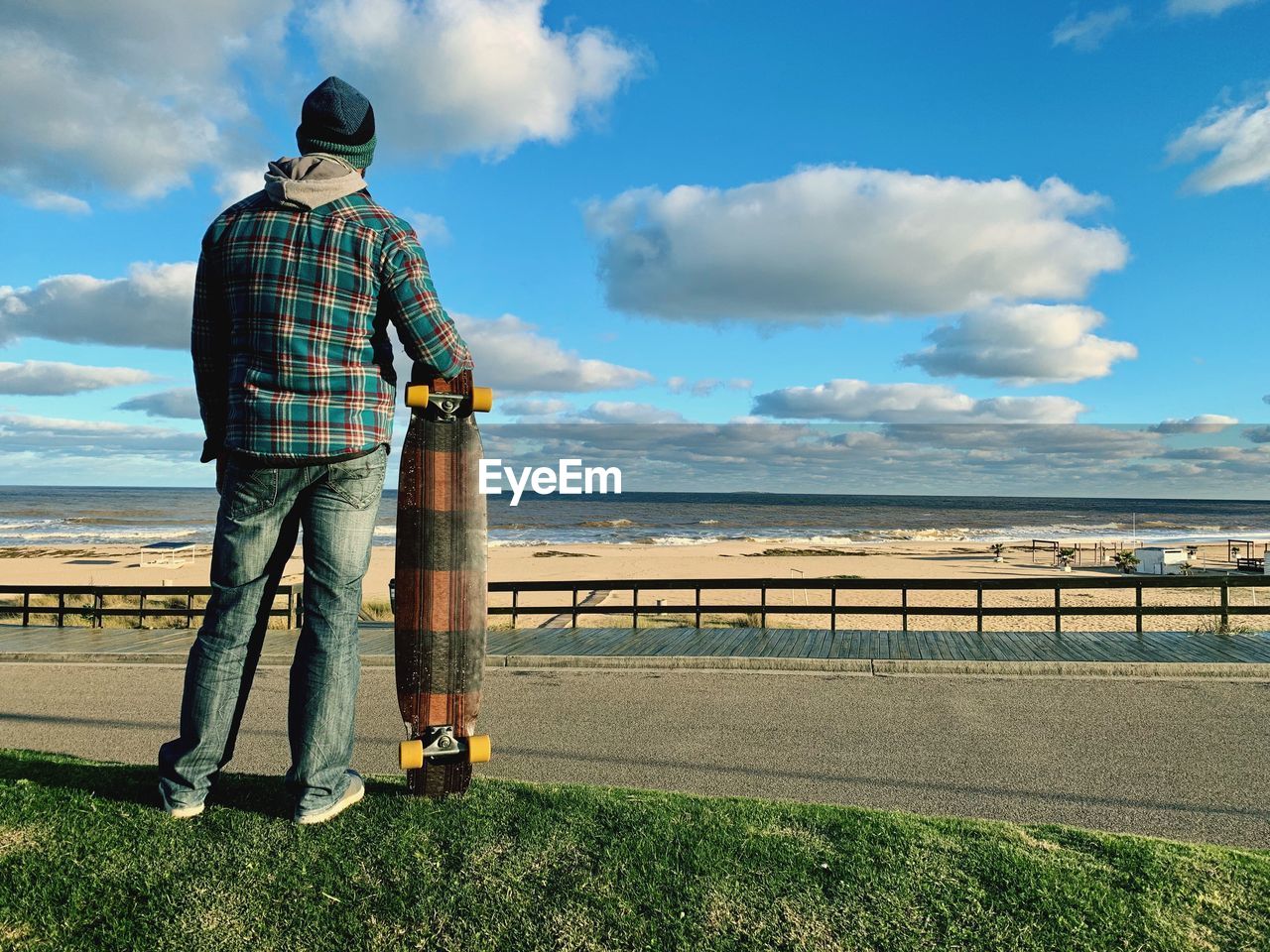 Rear view of man holding skateboard while looking at sea against sky