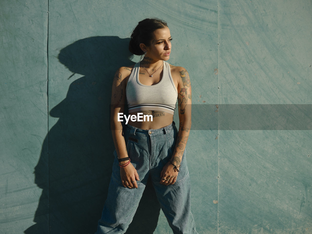 Confident young female with tattoos wearing stylish outfit standing near wall in city and looking away