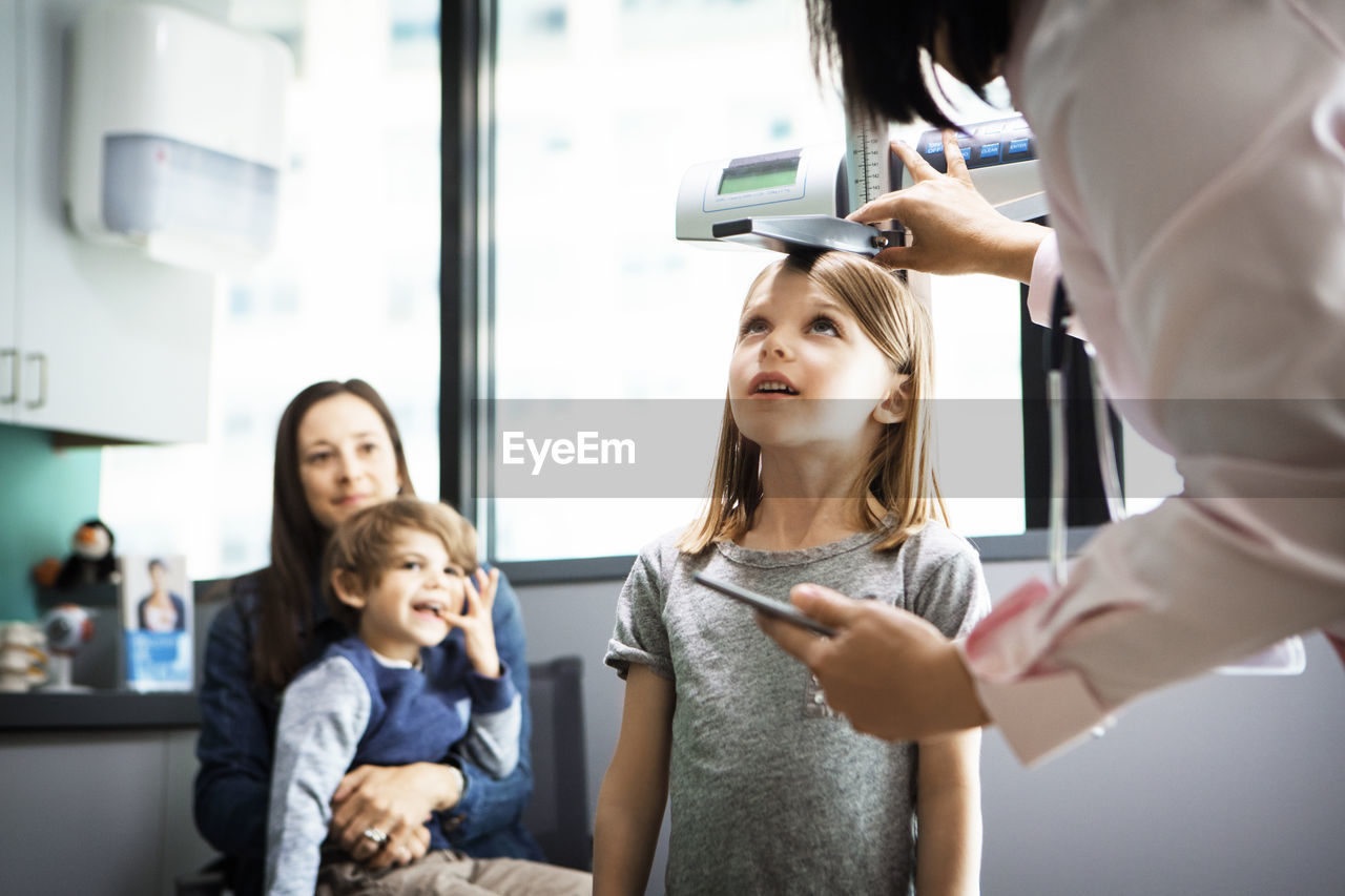 Doctor measuring girl's height while family sitting at clinic