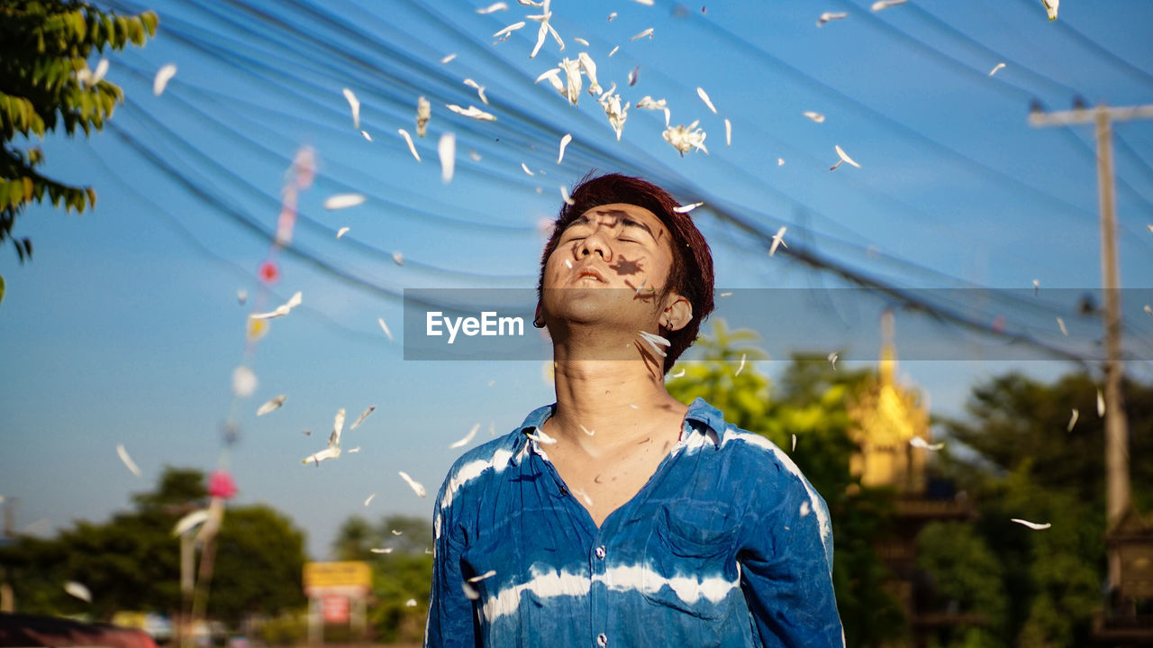 Petals falling on young man standing against blue sky