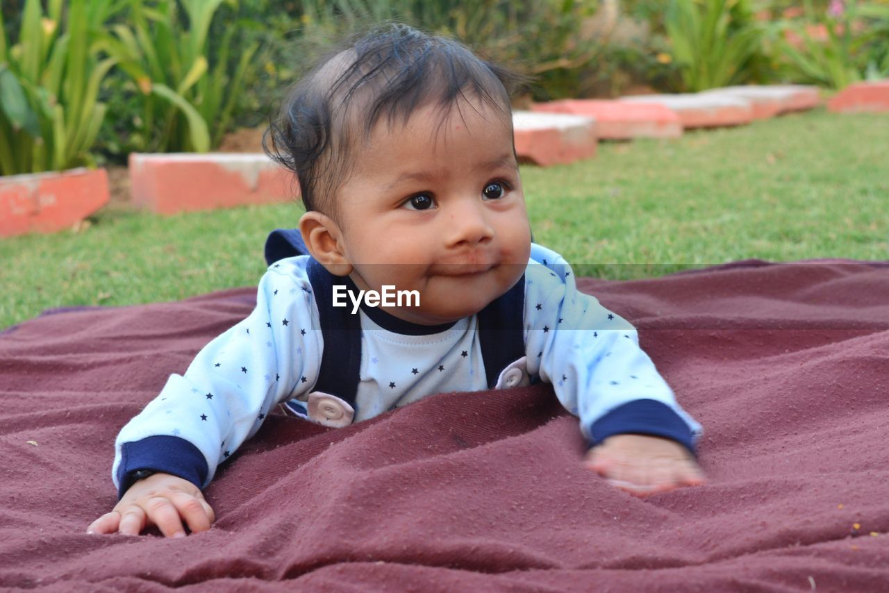 Cute baby boy looking away while lying on blanket in lawn