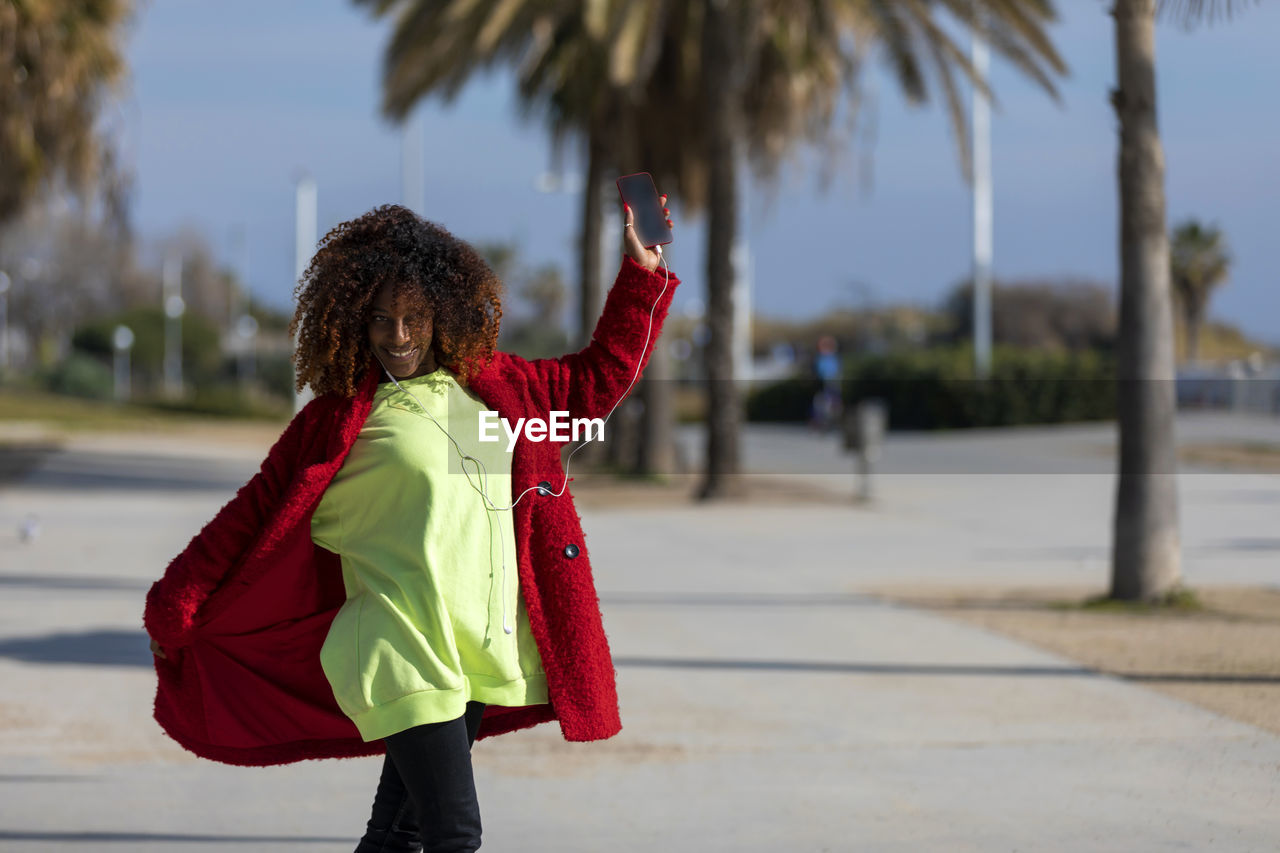 Portrait of woman dancing while listening music on mobile phone against trees