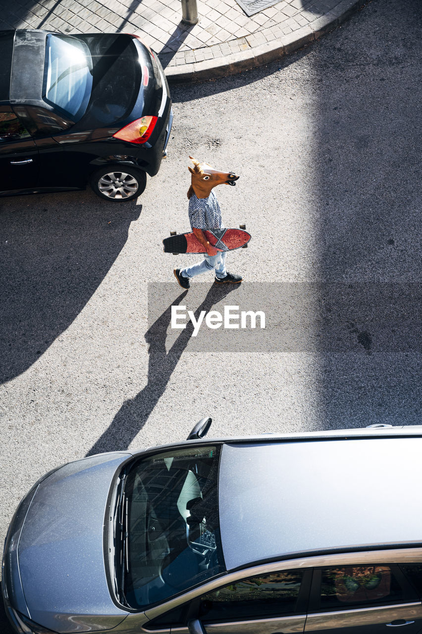 Mid adult man wearing horse mask holding skateboard while walking amidst cars on street in city