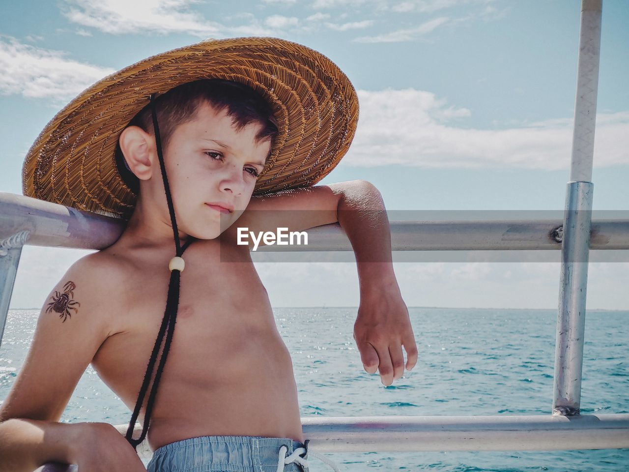 Thoughtful shirtless boy wearing hat while standing by sea against sky