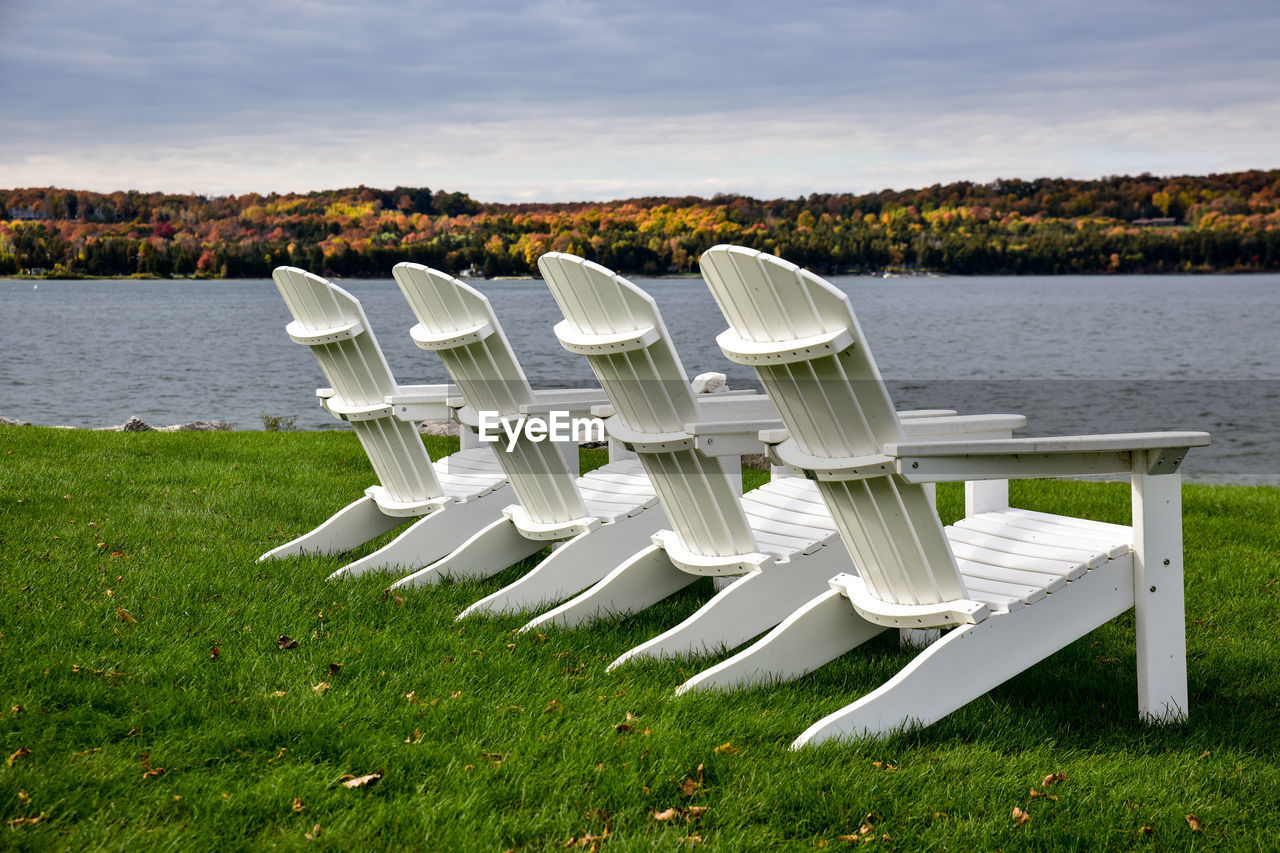 Empty adirondack chairs at grassy lakeshore against cloudy sky