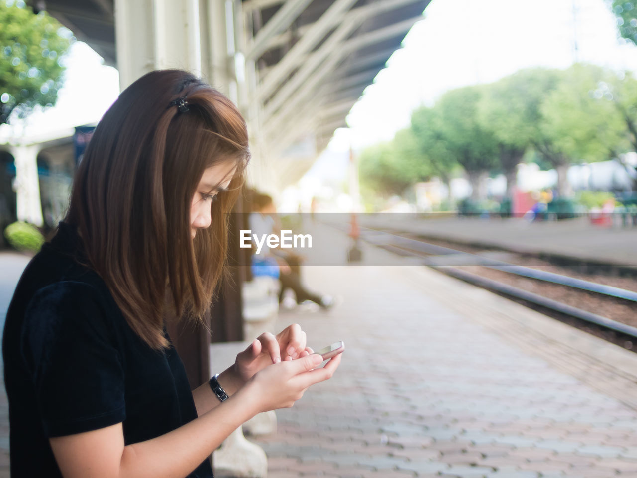 Side view of young woman using phone while sitting on railroad station
