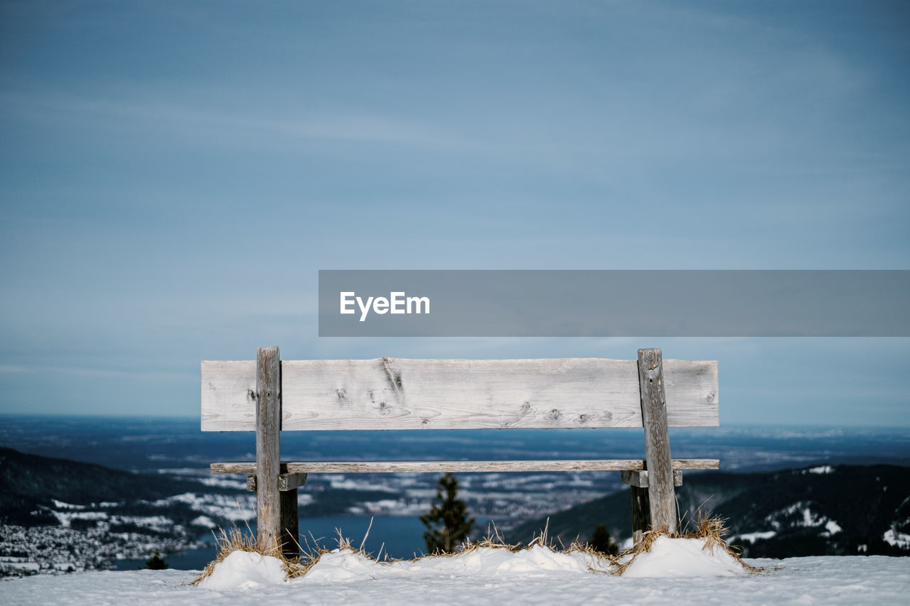 Wooden bench on snow covered land against sky