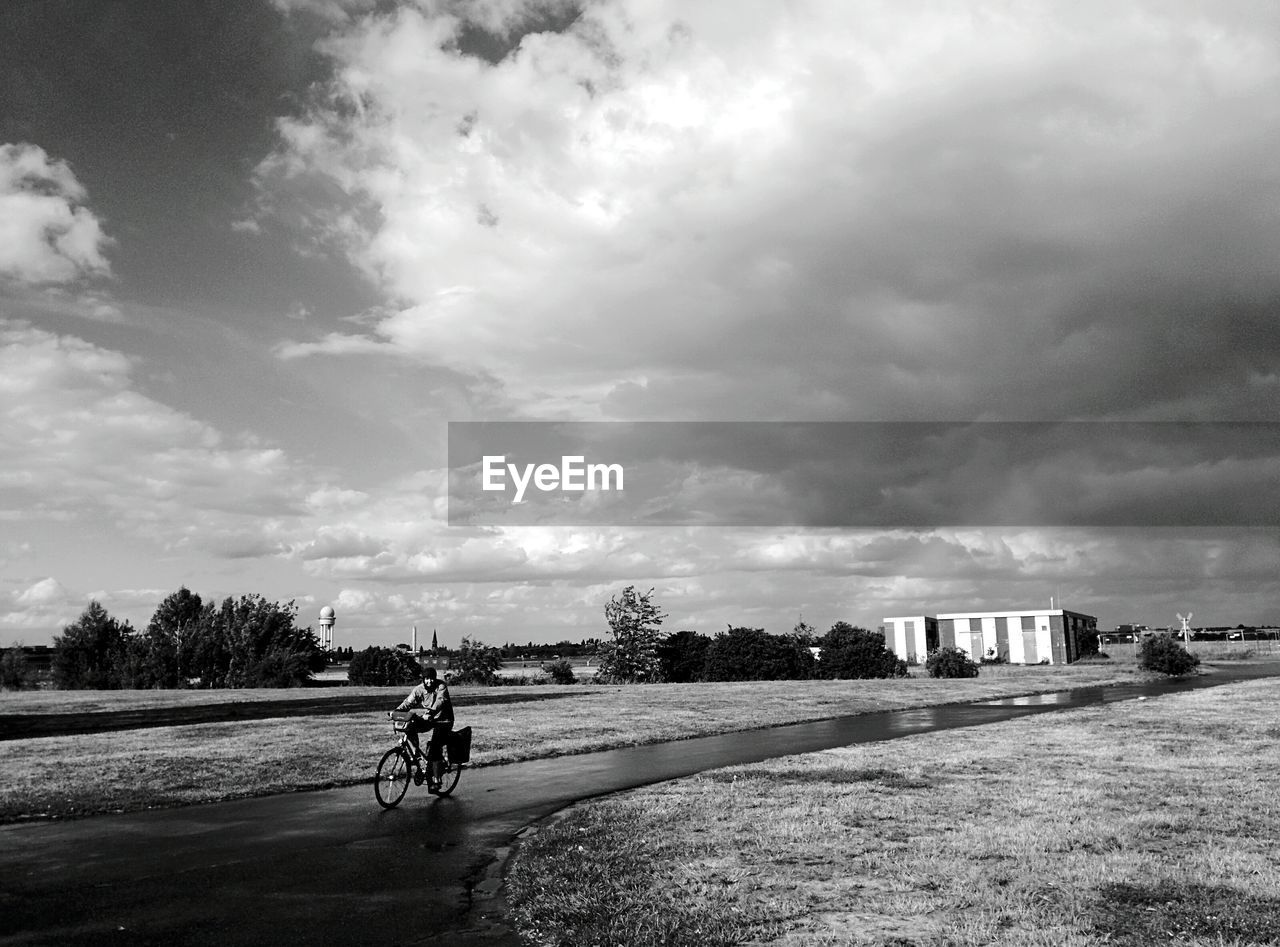 Man cycling on road amidst field against cloudy sky