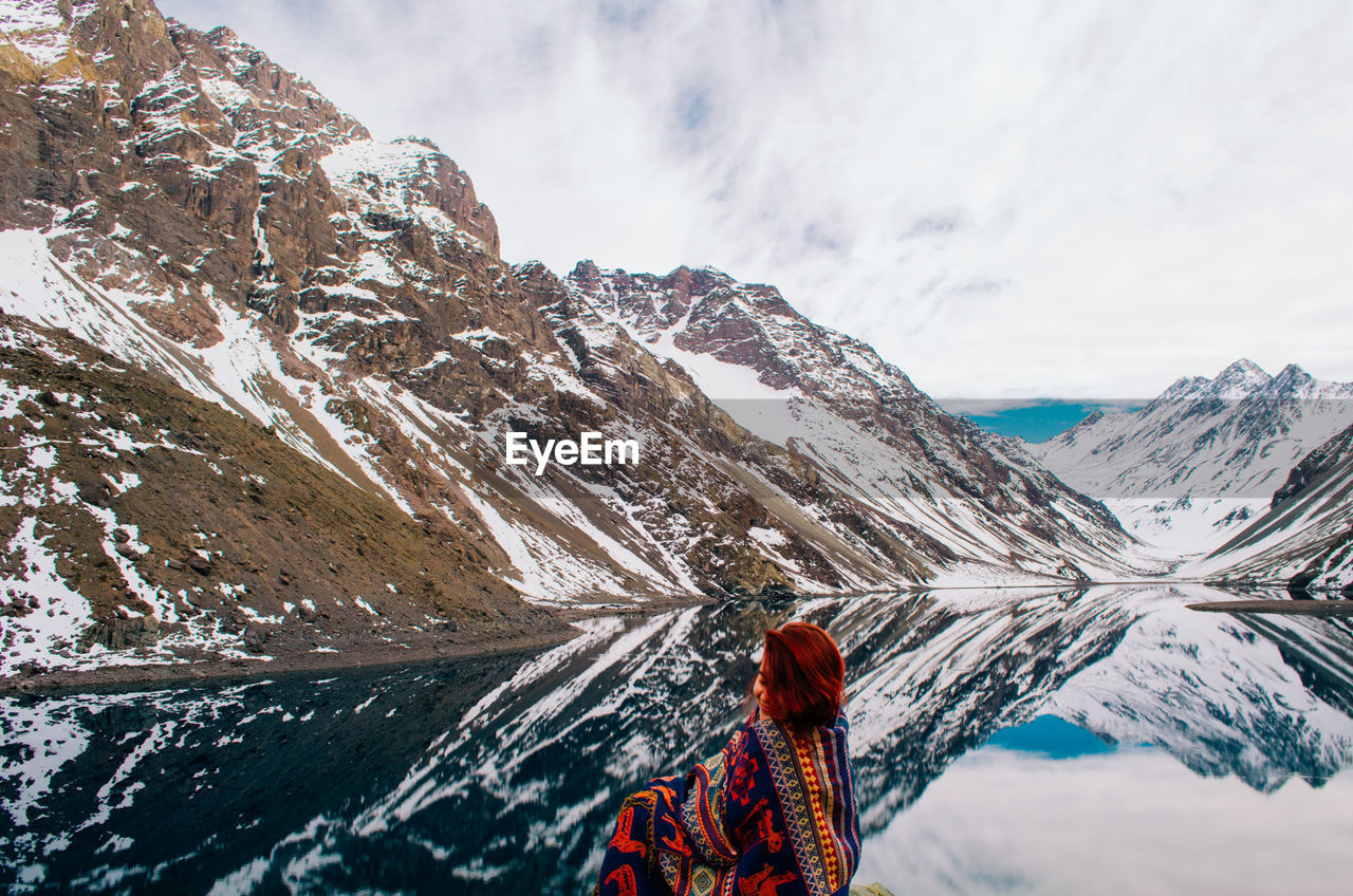 Rear view of young woman sitting by lake against mountain range