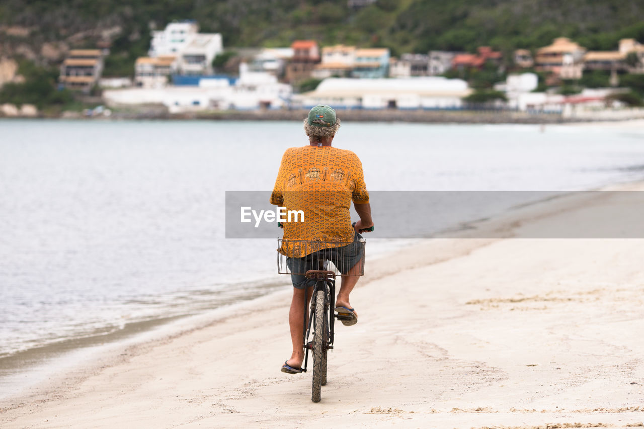 REAR VIEW OF MAN RIDING BICYCLE ON SEA SHORE
