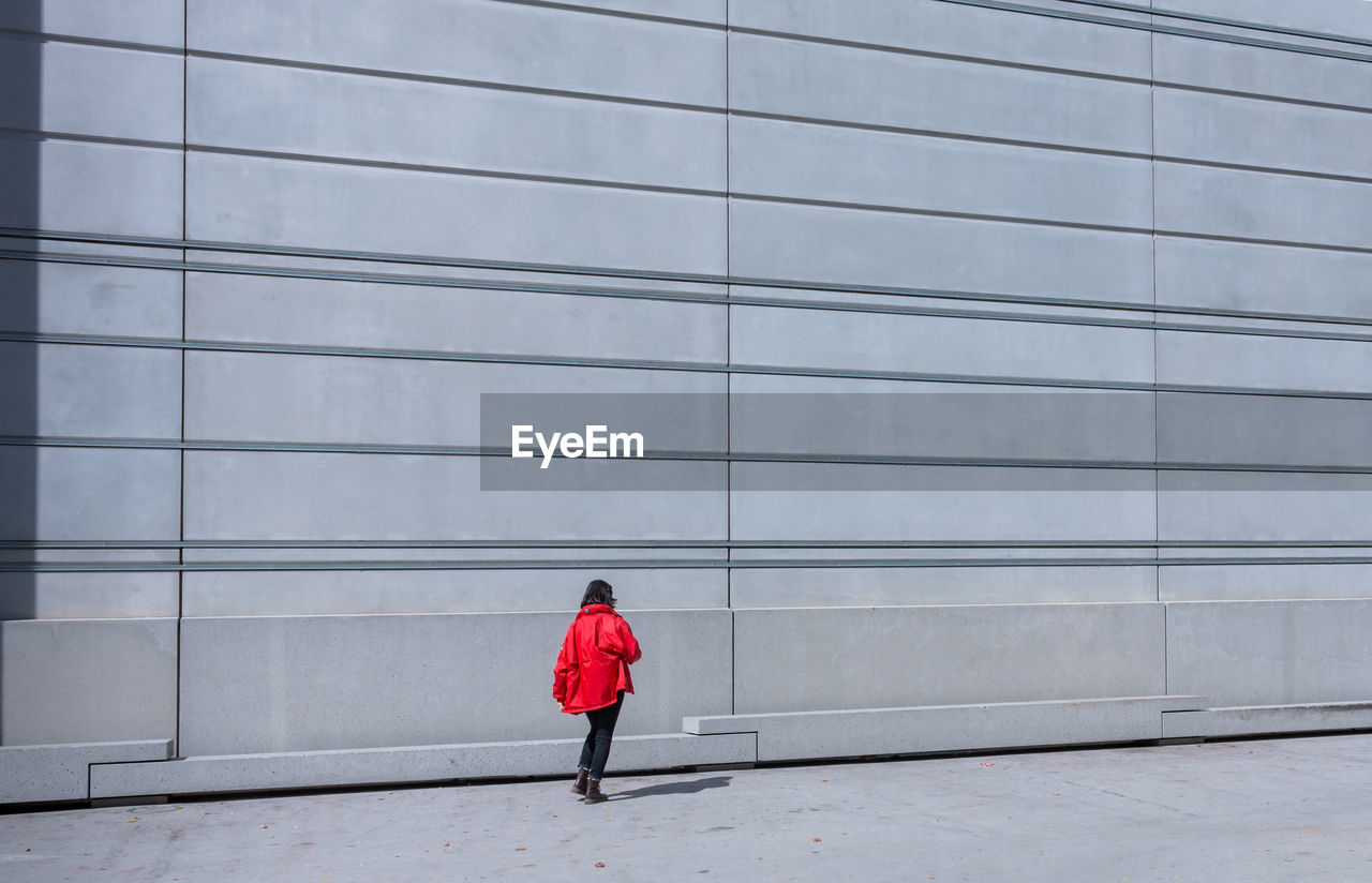 A young woman in a red jacket walks along a windowless grey façade with a pattern of lines