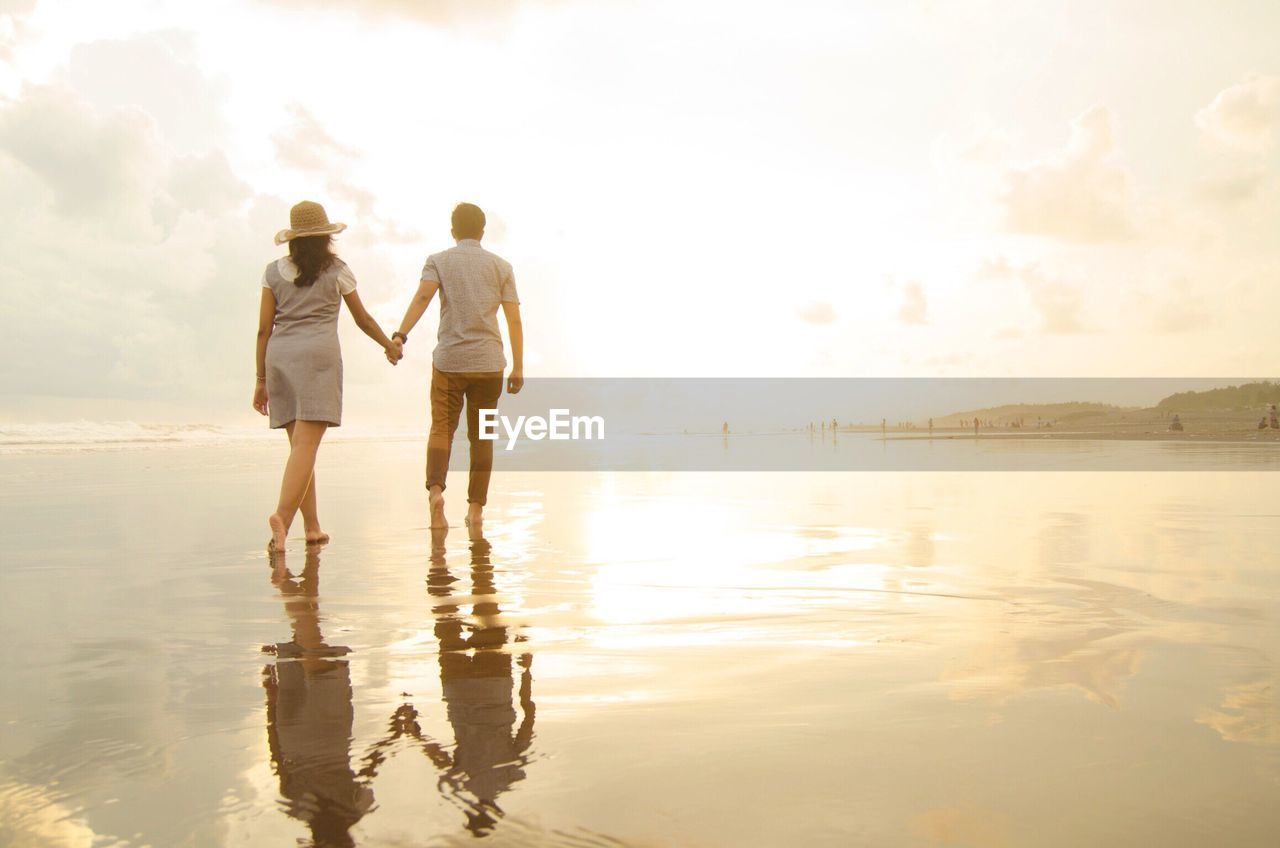 Rear view of couple walking at beach against cloudy sky during sunset