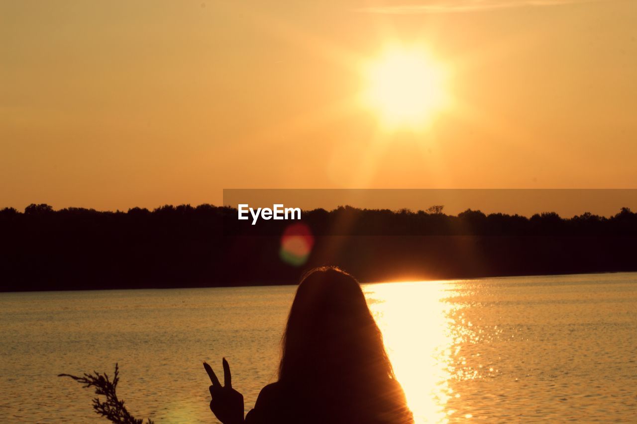 Silhouette woman showing peace sign by lake against clear sky during sunset
