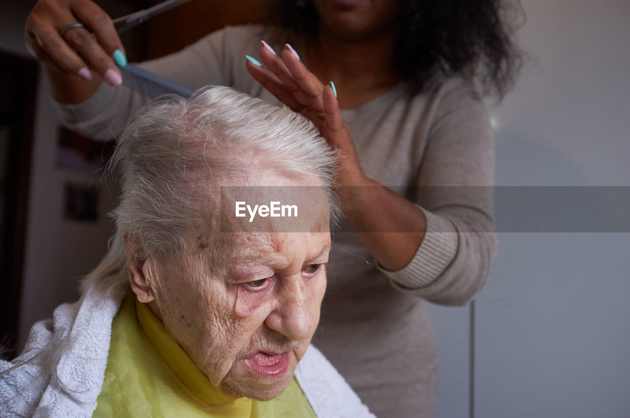 Caring african american woman caregiver, cutting her elderly woman hair at home