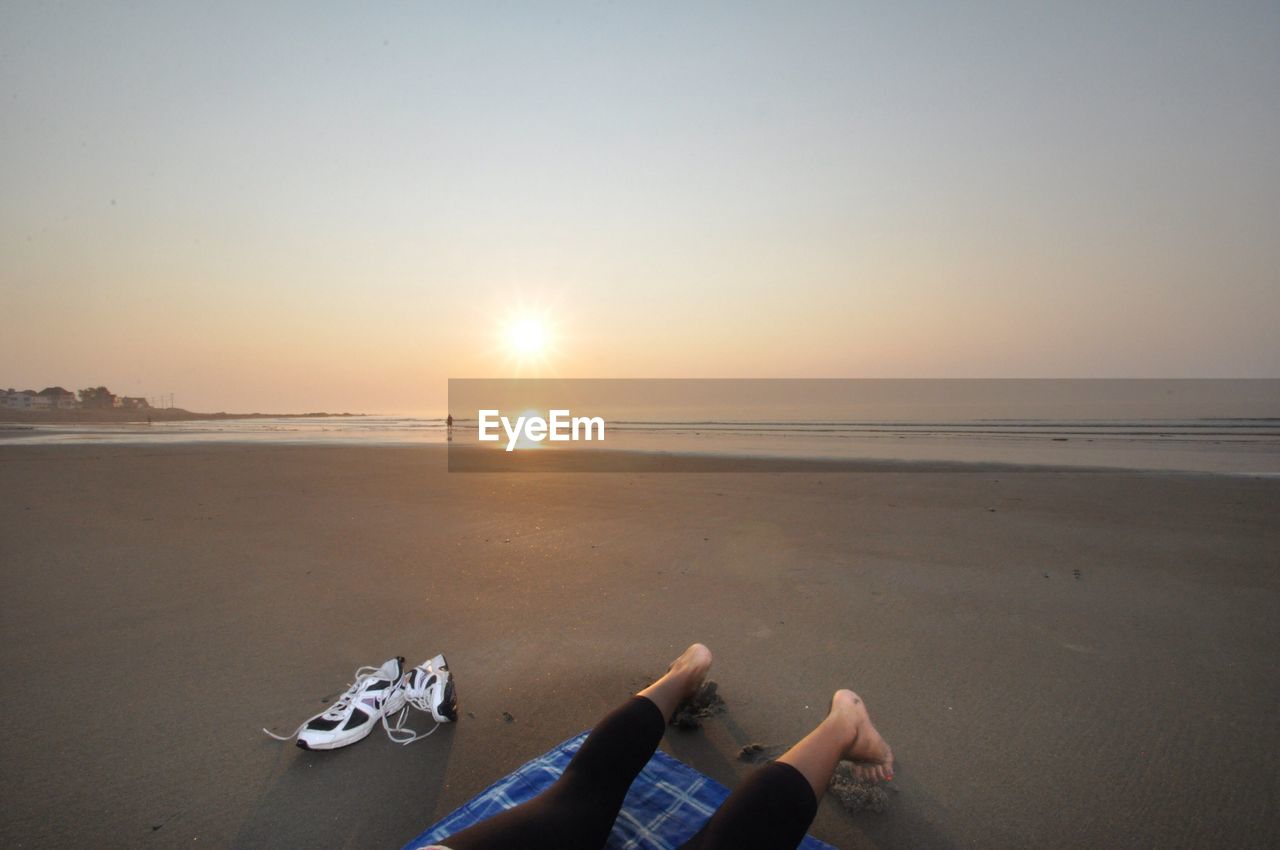 Low section of woman relaxing at beach against sky during sunset