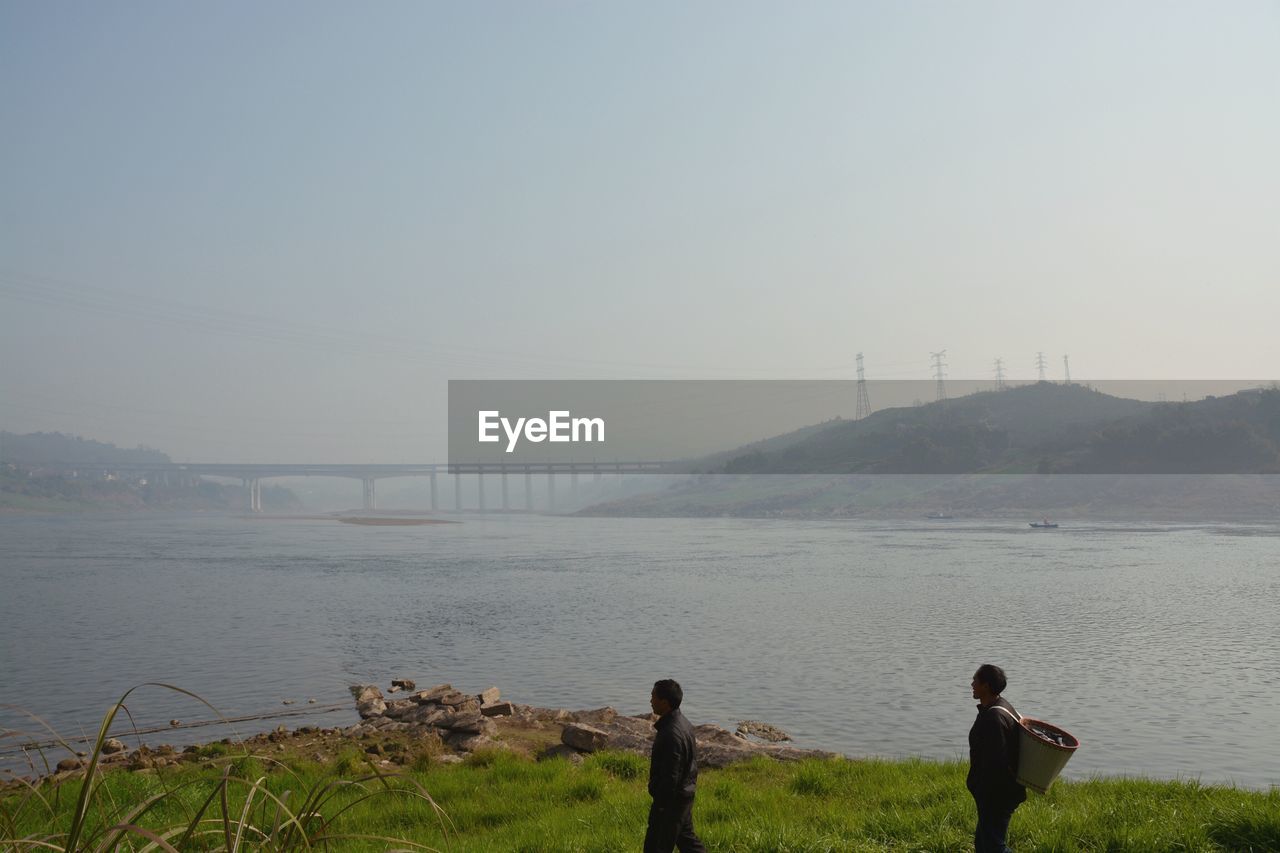 People standing on grass by river against sky